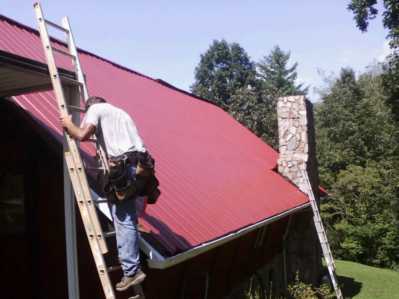 Roofer on a ladder beside a red metal roof and stone chimney — Soffit & Fascia Repair by Roofing Brothers in Marathon